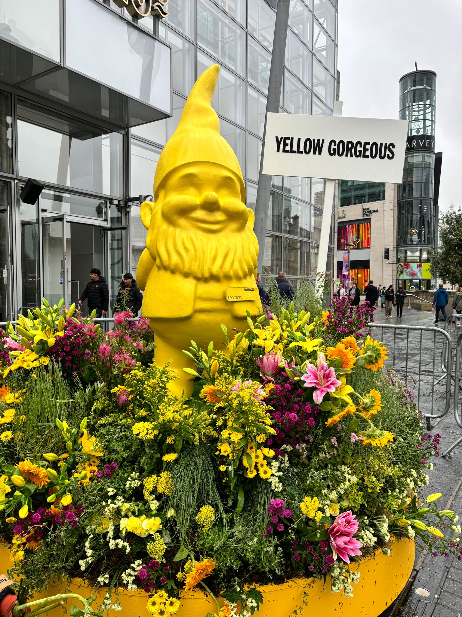 Yellow Gnome for Manchester Flower Festival. Yellow gnome surrounded by a garden of yellow and pink flowers like achillea, sunflowers, lilies, banksia and status. Outside Selfridges in Manchesrter.