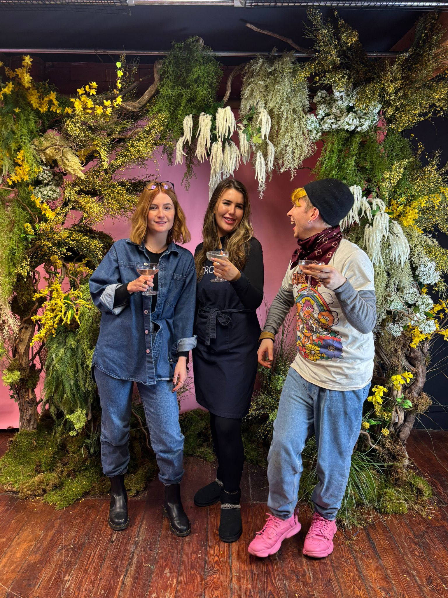 Installation workshop at Frog Flower School in Manchester Northern Quarter. Three people stand in front of a floral arch of seasonal greenery and yellow flowers, with white amarnthus and ginestra. Installation includes moss. Flower Workshop.