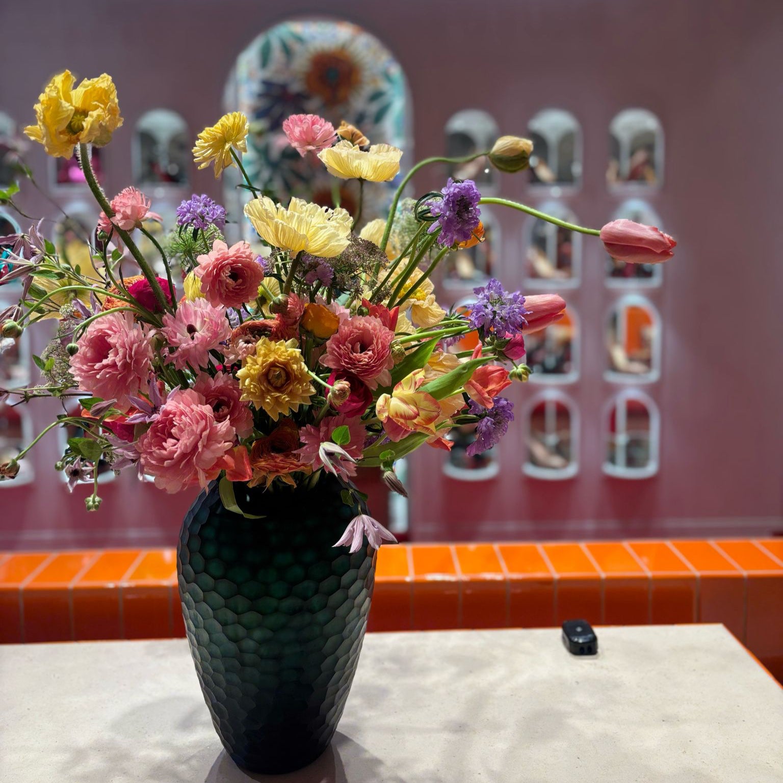 Seasonal Spring flower arrangement for corporate flower installation at Christian Louboutin in Selfridges Exchange Square. Vase of yellow poppies, pink ranunculus, french tulips, clematis, lilac scabiosa and butterfly ranunculus.