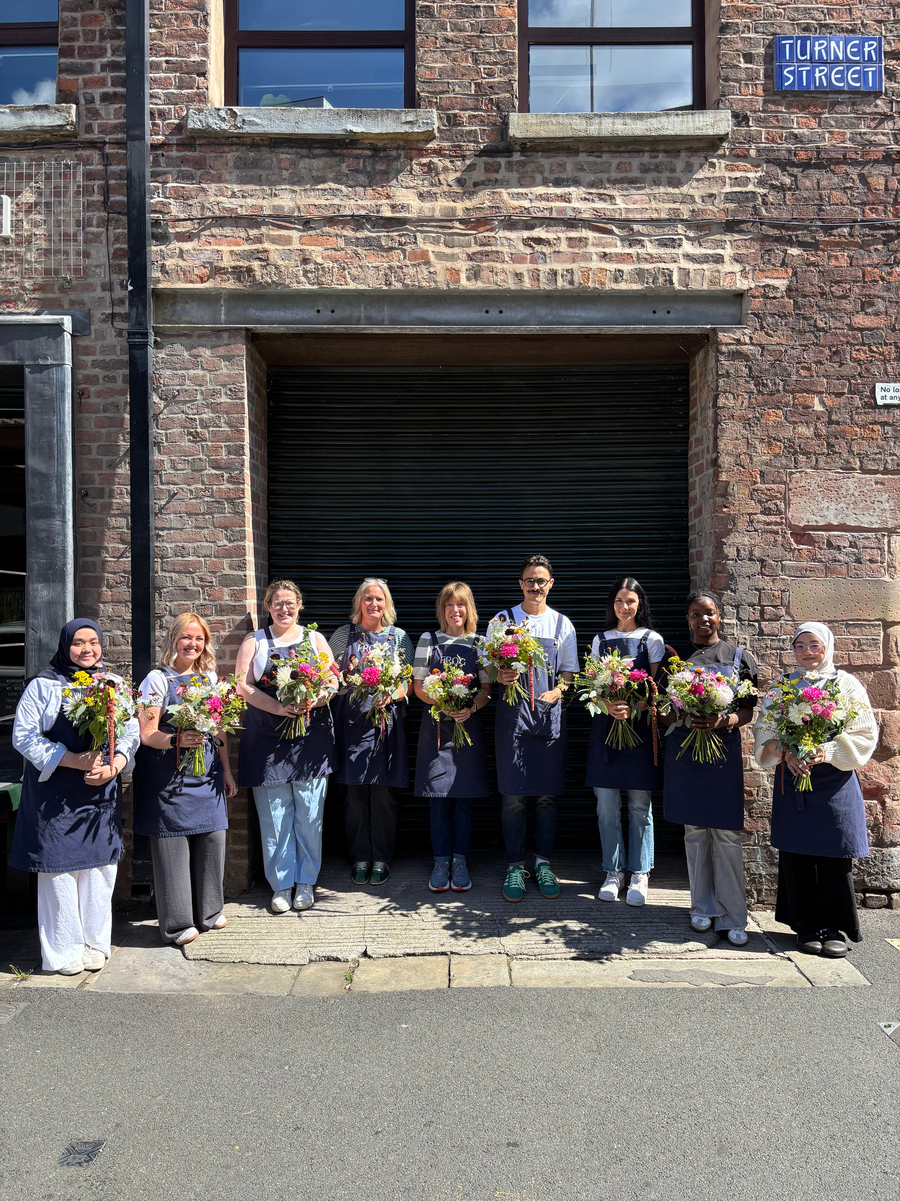 Group of people holding flower bouquets in front of a brick building with 'Turner Street' sign.
