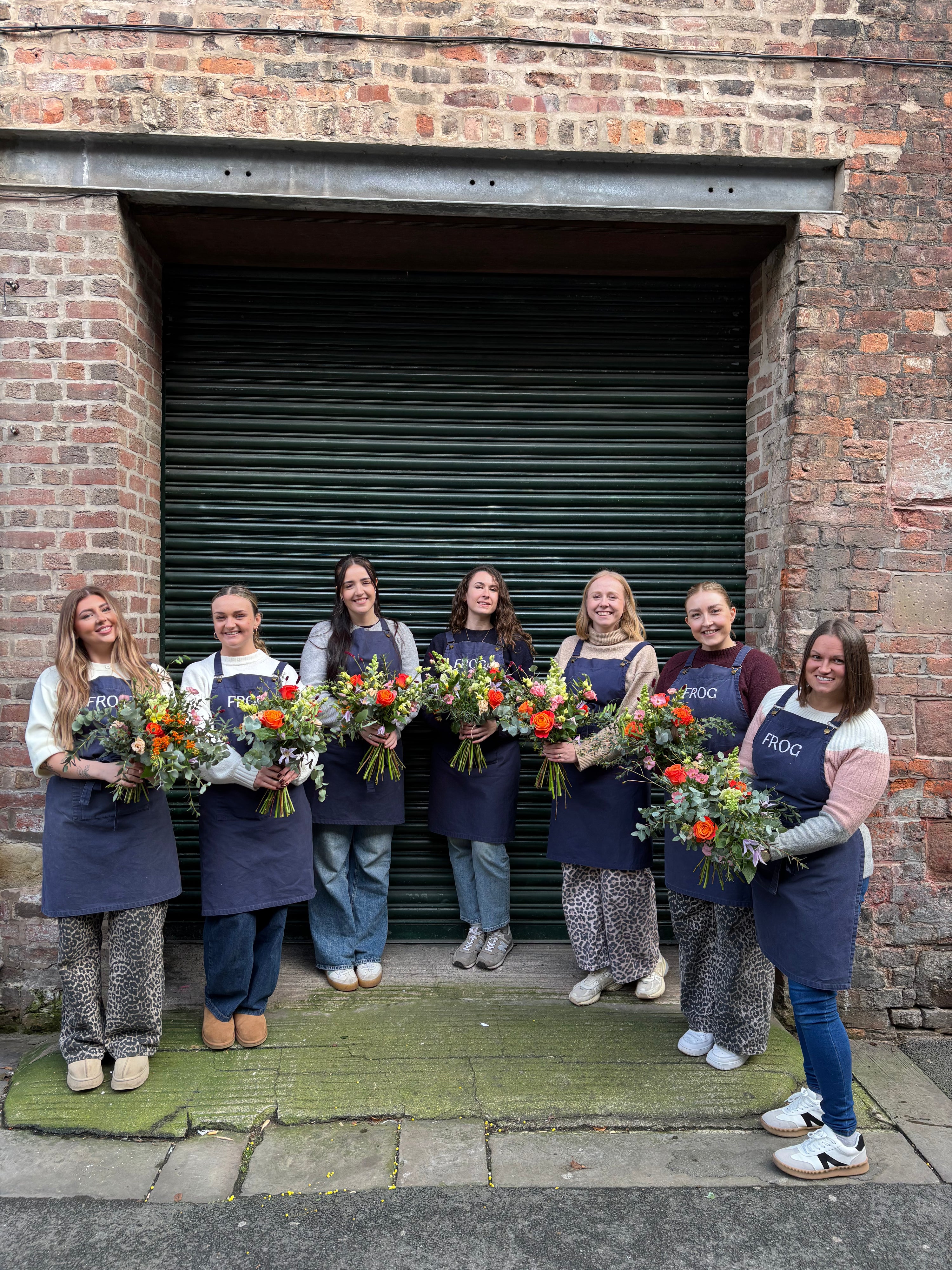 Group of women holding floral arrangements in front of a brick building with a green shutter.