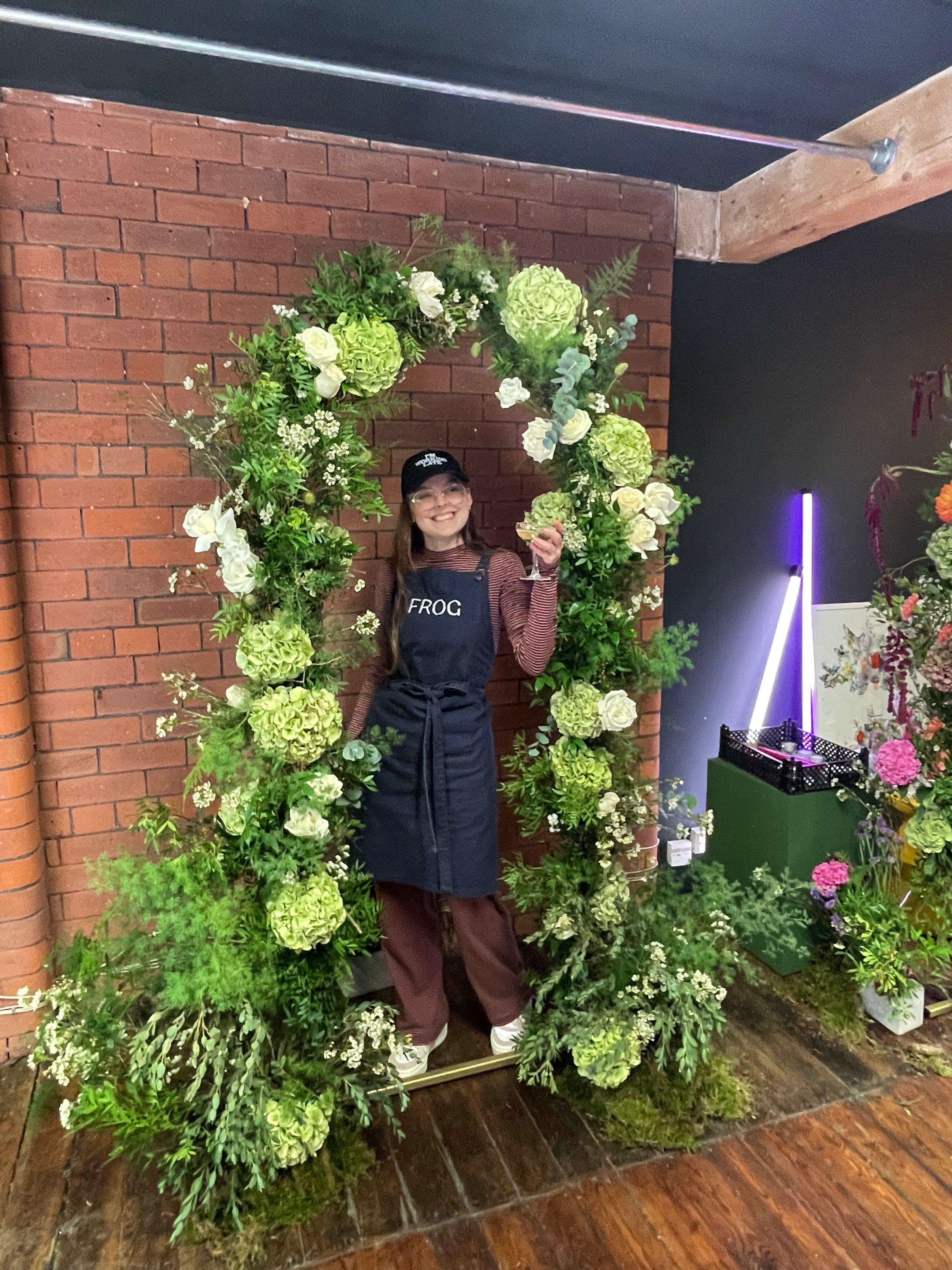 Person standing behind a floral archway with a brick wall and DJ setup in the background
