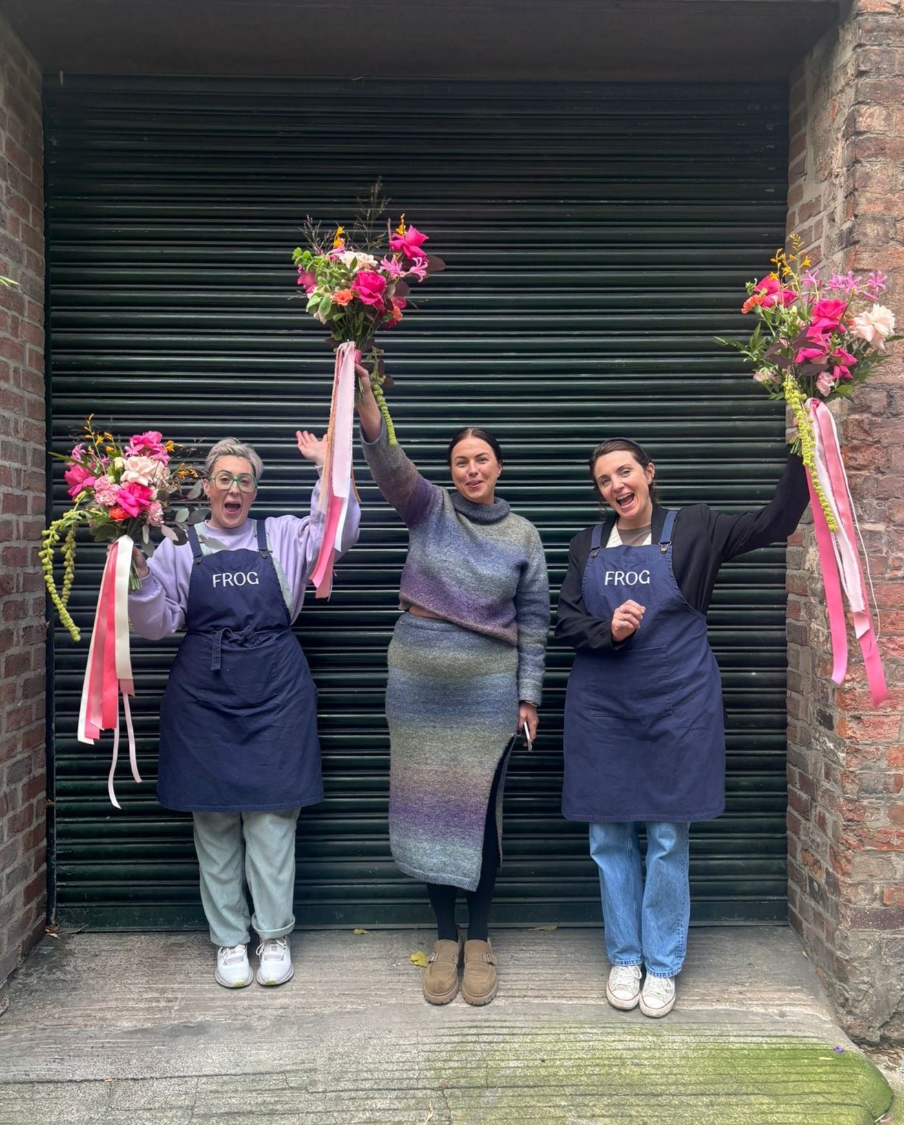 Three individuals participating in a wedding flower arrangement course, each holding a different floral arrangement, standing in front of a black shutter.