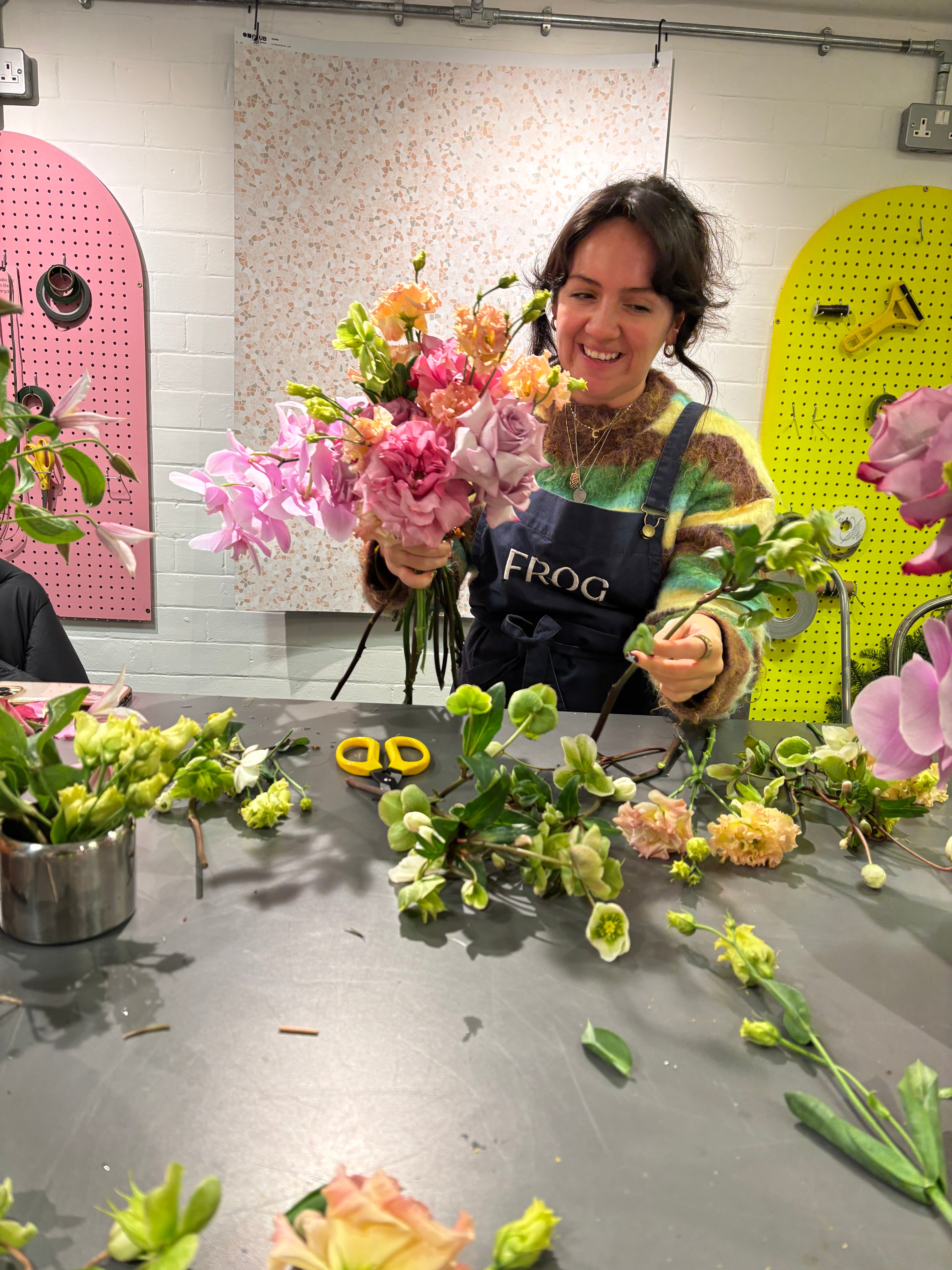 Woman arranging flowers in a workshop setting with colorful pegboards in the background.