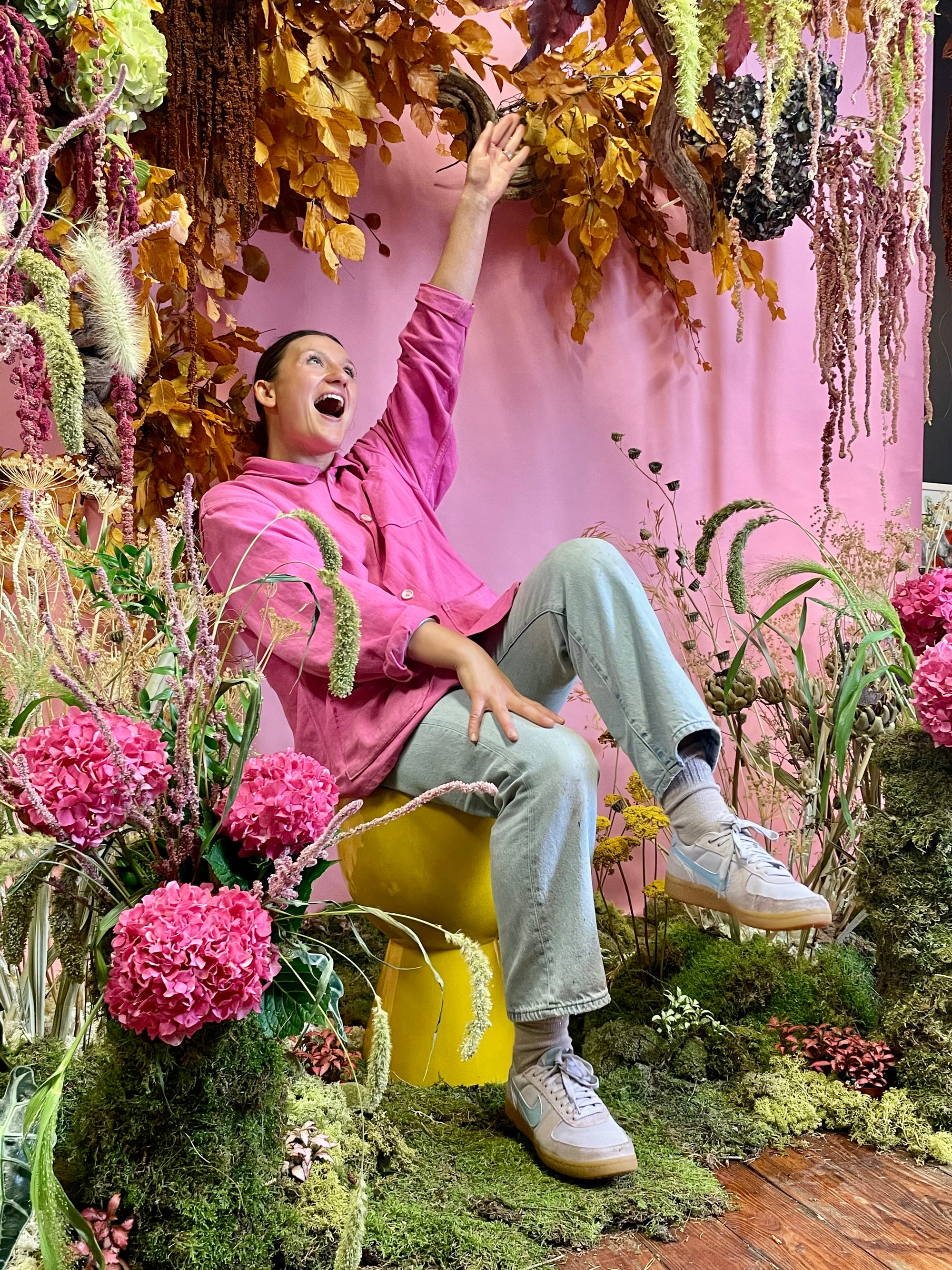 Person in a pink shirt sitting on a yellow stool surrounded by flowers and plants with a pink curtain in the background.