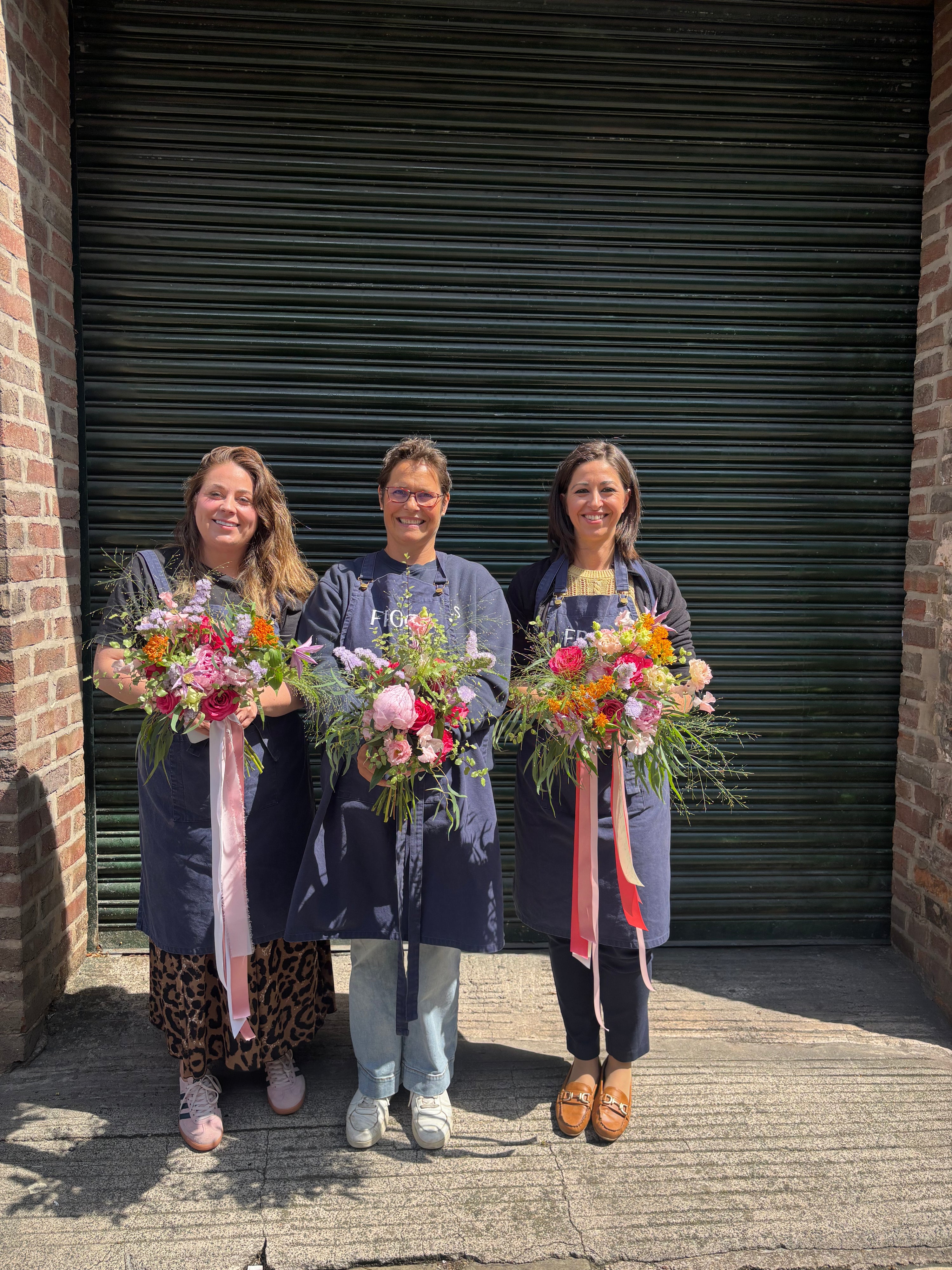 Three people holding floral bouquets in front of a brick building with a green shutter.