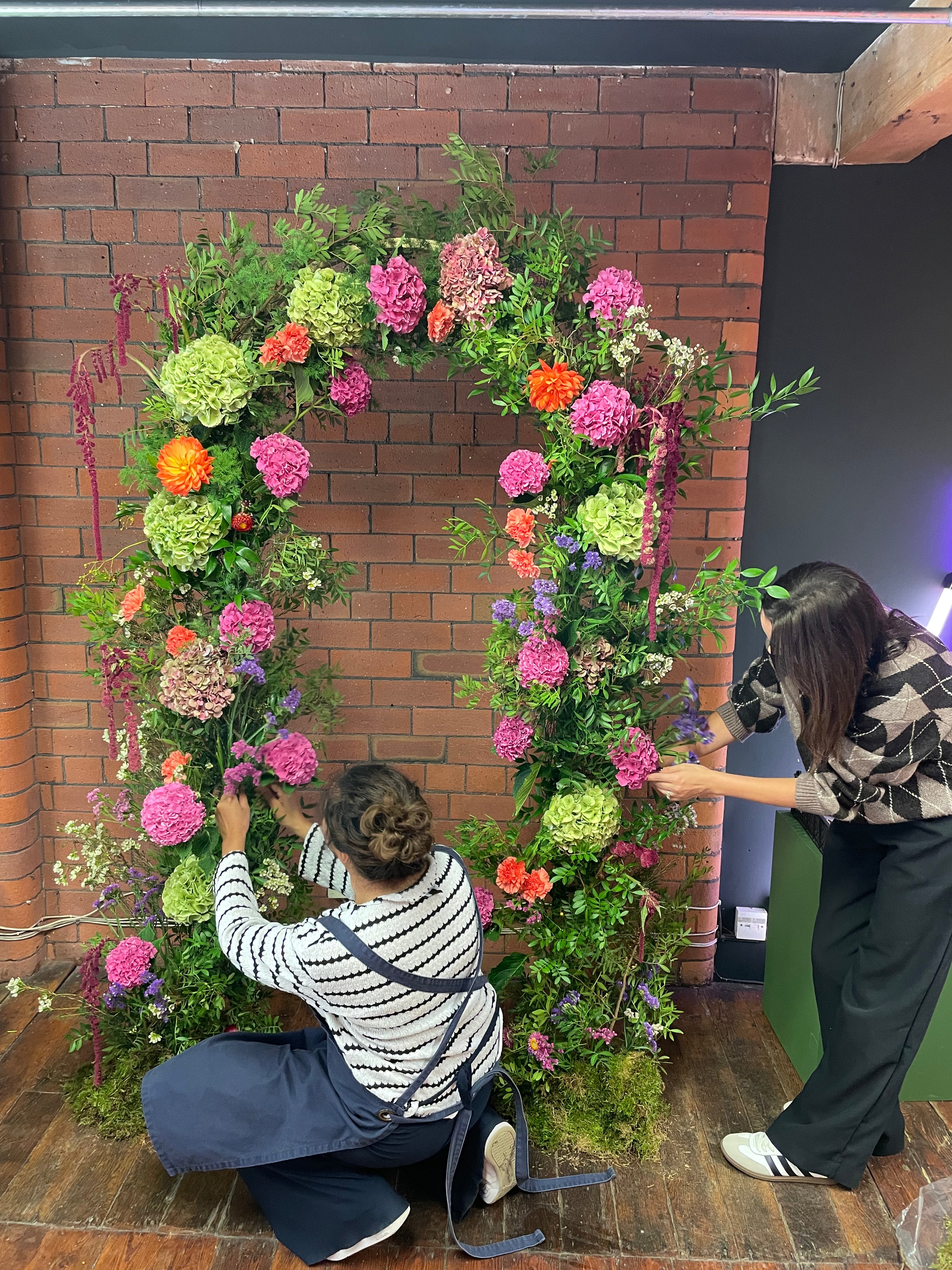 Two people arranging flowers in a large floral arch against a brick wall.