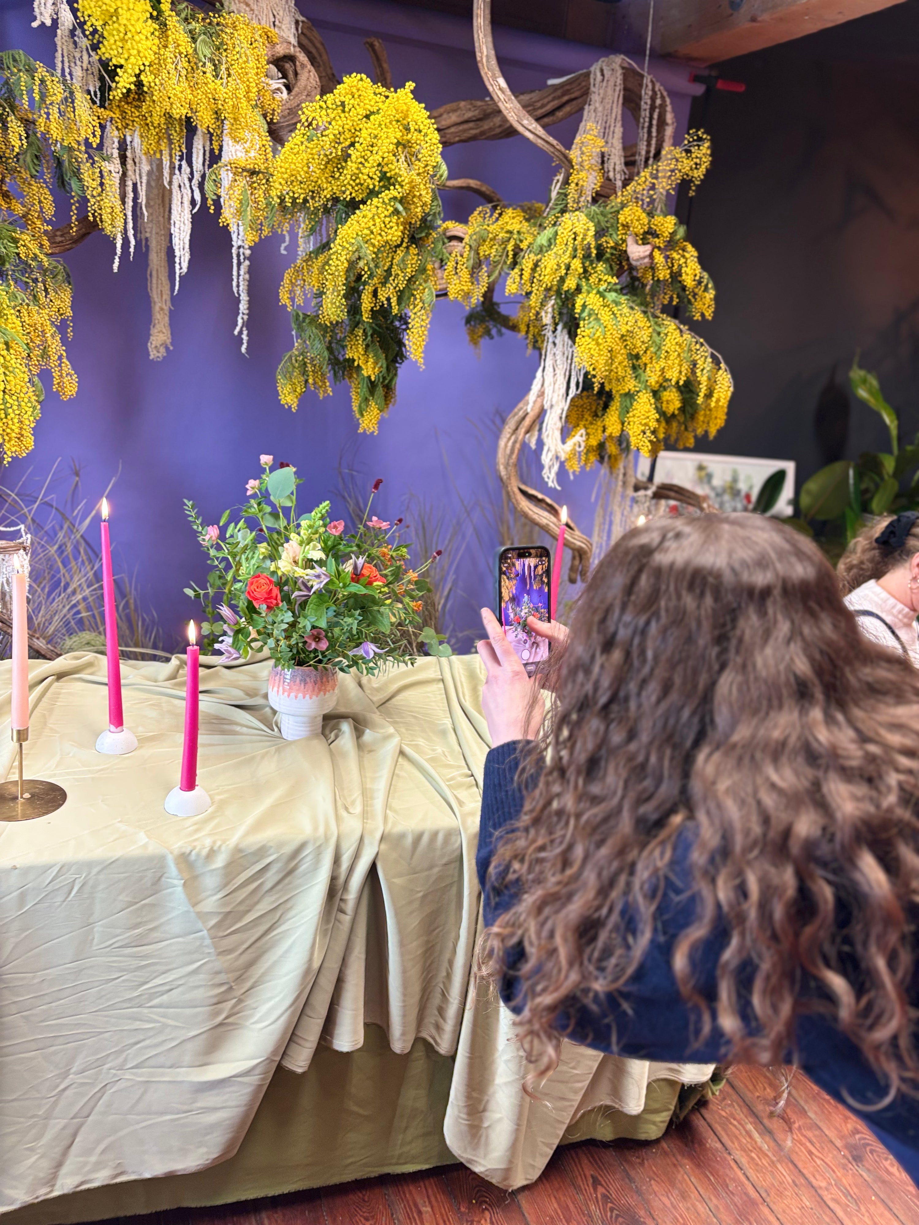 Person taking a photo of a decorated table with flowers and candles, against a purple wall with hanging yellow flowers.