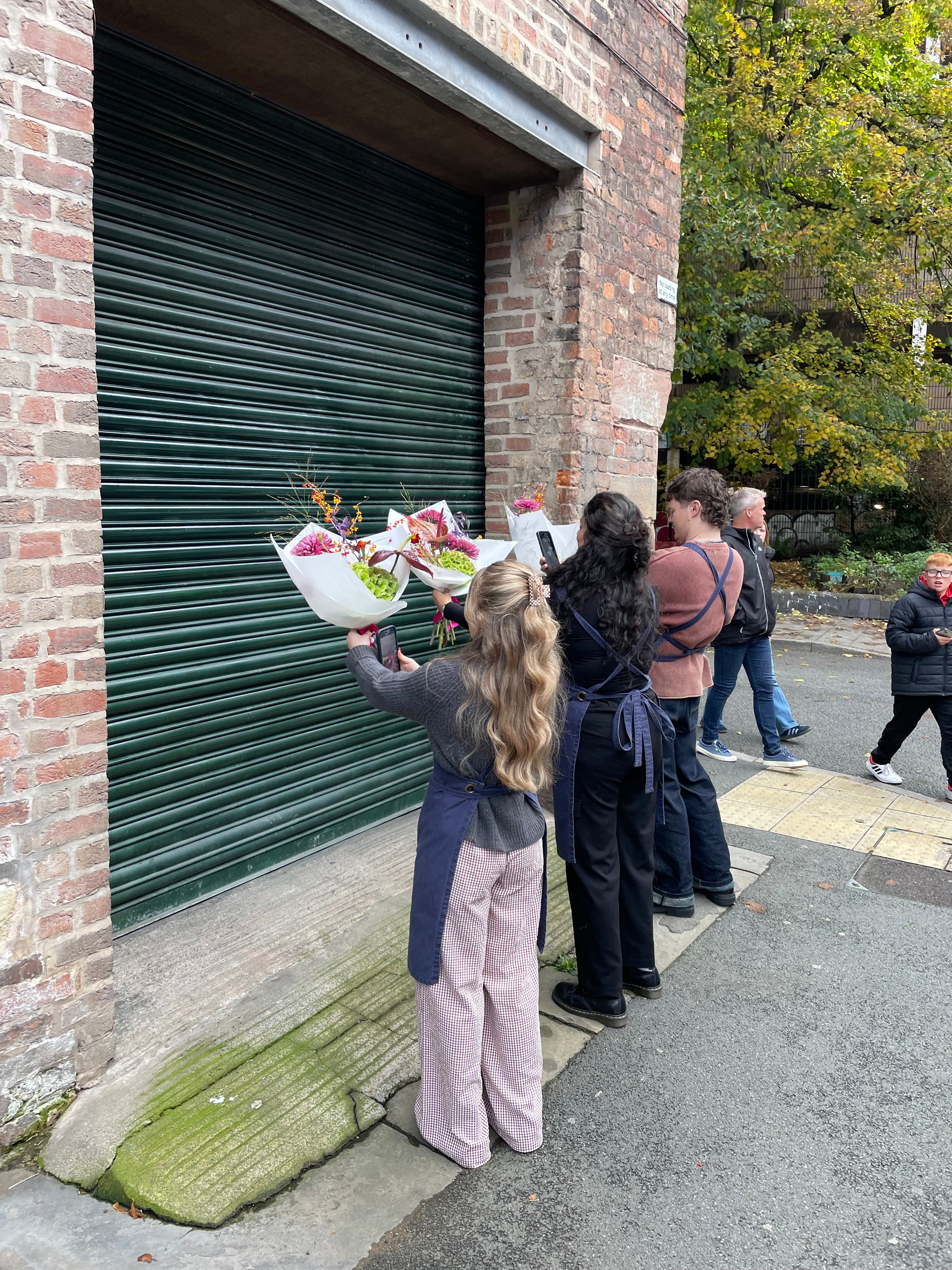 Group of people standing outside a building with a green shutter, holding flowers and a bouquet.