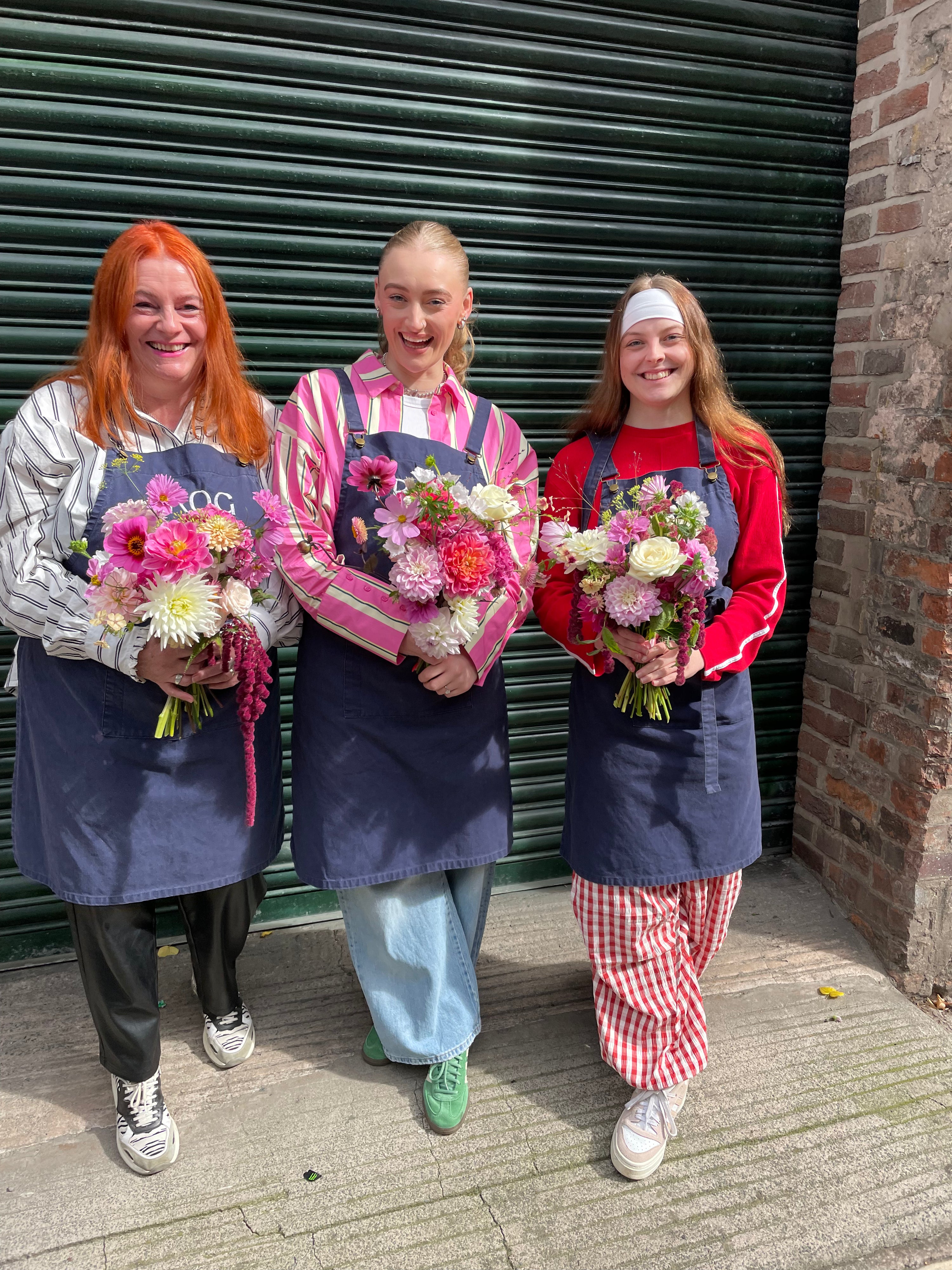 Three women holding flower bouquets in front of a closed shutter.